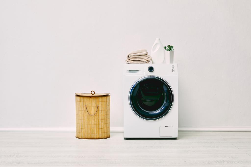 botella de detergente, toallas y planta en la lavadora moderna cerca de la cesta de lavandería en el baño
  - Foto, imagen