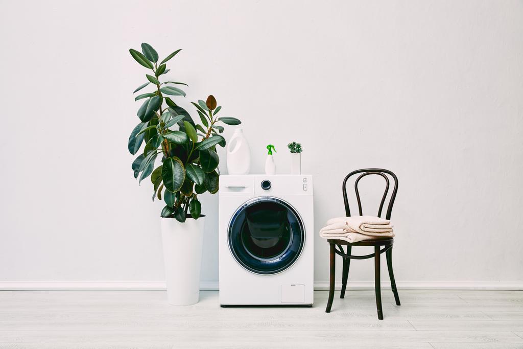 grüne Pflanzen in der Nähe von Flaschen auf Waschmaschine in der Nähe von Stuhl mit Handtüchern  - Foto, Bild