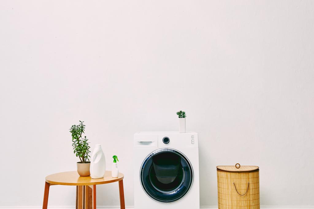 plantas verdes y botellas cerca de mesa de centro de madera, cesta de lavandería y lavadora moderna en el baño
  - Foto, imagen