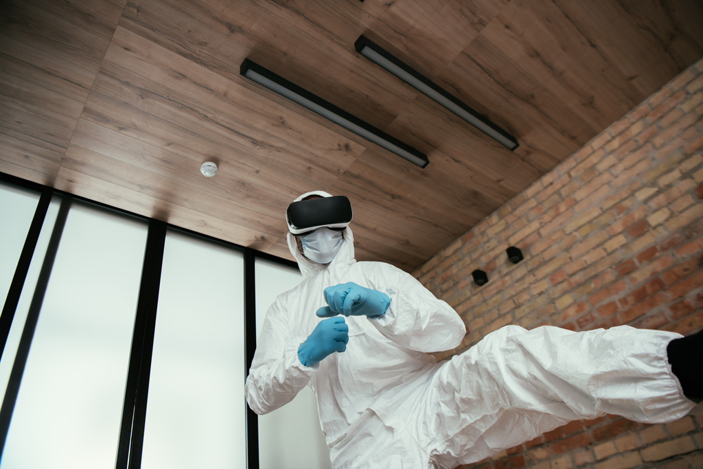 low angle view of man in hazmat suit, medical mask, latex gloves and virtual reality headset gaming in living room  - Photo, Image