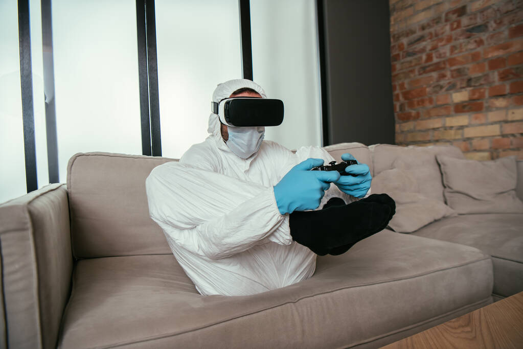 KYIV, UCRANIA - 11 DE ABRIL DE 2020: hombre con traje de materiales peligrosos, máscara médica, guantes de látex y auriculares de realidad virtual jugando videojuegos en el sofá
 - Foto, imagen
