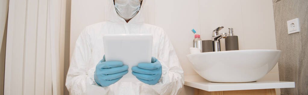 colpo panoramico di uomo in tuta hazmat, maschera medica e guanti in lattice utilizzando tablet digitale in bagno
  - Foto, immagini