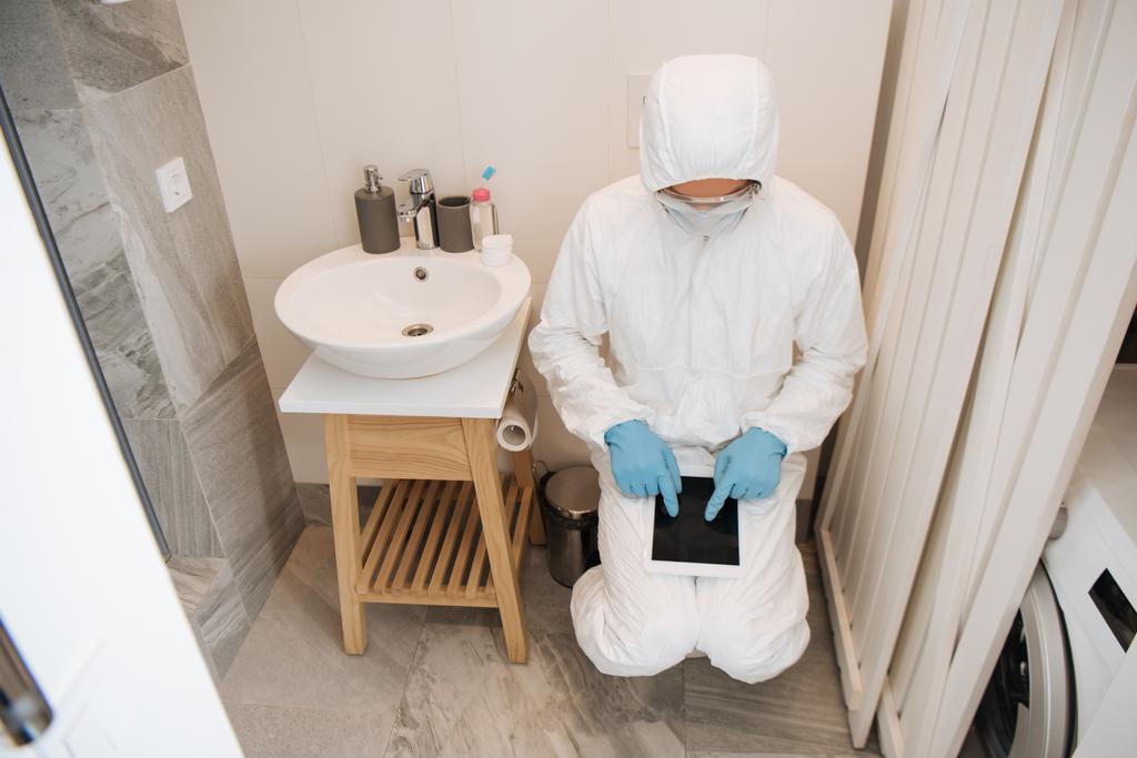 muž v ochranném obleku, lékařské masce, latexových rukavicích a brýlích ukazujících prsty na digitální tablet s prázdnou obrazovkou v koupelně  - Fotografie, Obrázek