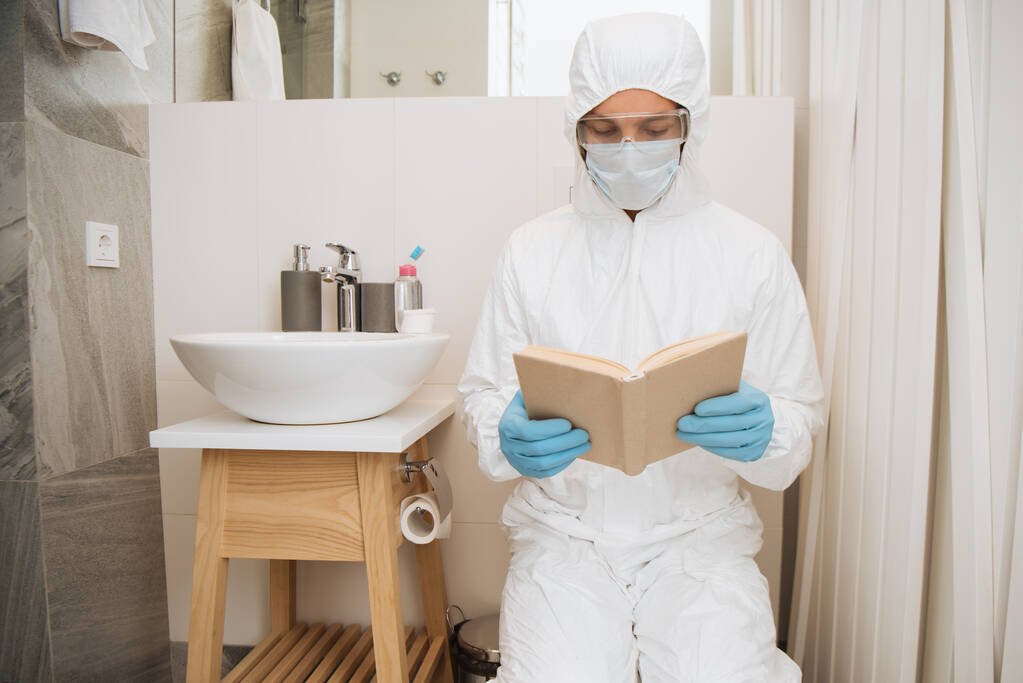 Mann im Warnanzug, medizinischer Maske, Latexhandschuhen und Brille beim Lesen von Büchern im Badezimmer  - Foto, Bild