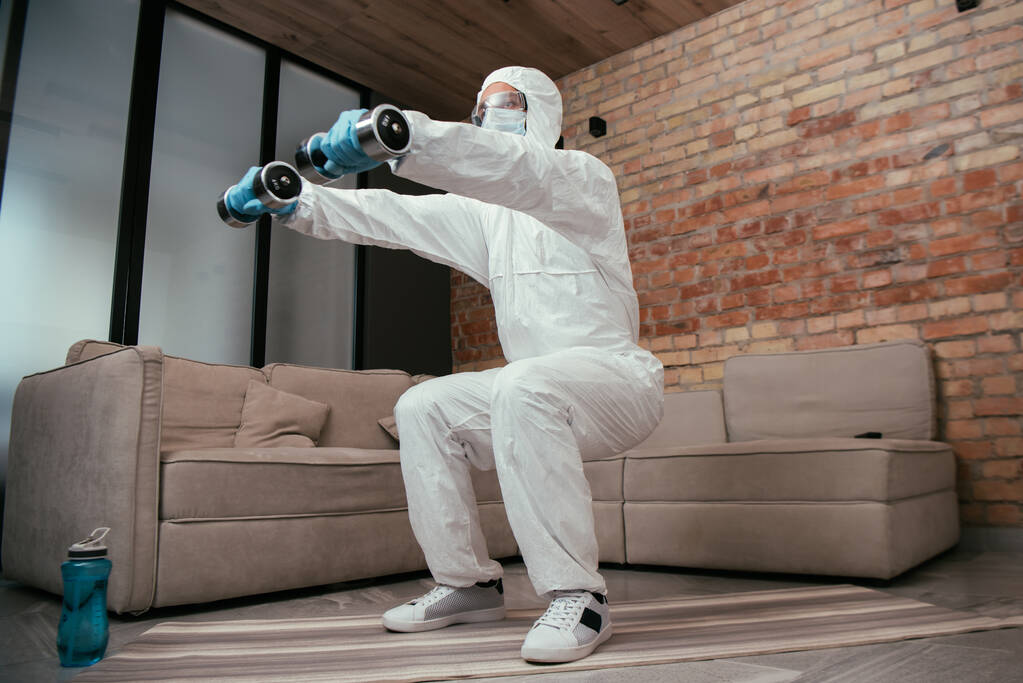 sportos férfi vegyvédelmi ruhában, orvosi maszk és szemüveg csinál guggolás gyakorlat súlyzókkal a nappaliban  - Fotó, kép