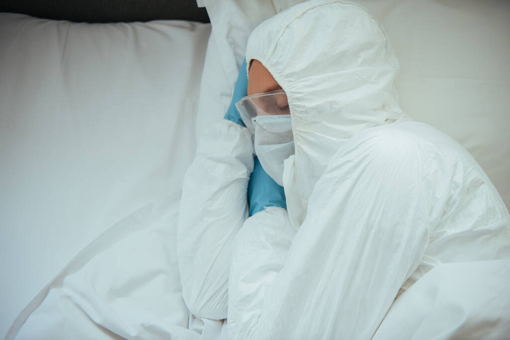 bovenaanzicht van de mens in hazmat pak, medisch masker en bril slapen op bed - Foto, afbeelding