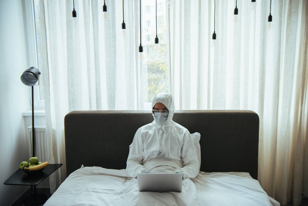 freelancer em terno hazmat, máscara médica e óculos usando laptop na cama
 - Foto, Imagem