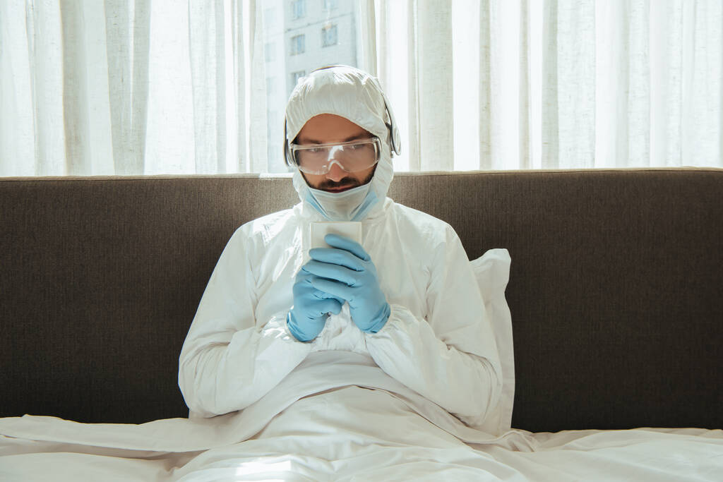 man in hazmat suit, headphones, medical mask, latex gloves and goggles holding cup with drink in bed - Photo, Image