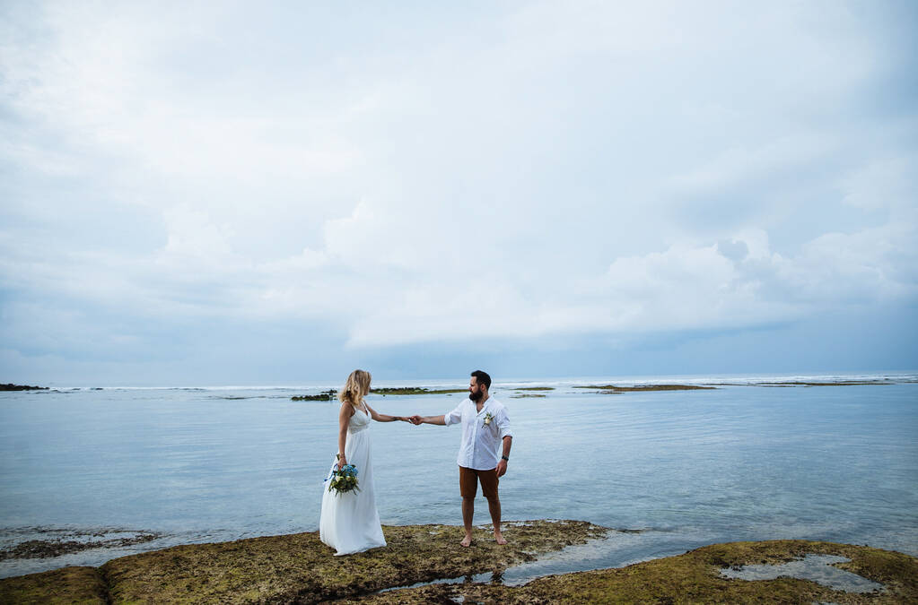 恋人同士-結婚式の日に花嫁と新郎が抱擁し、インドネシアのバリのエキゾチックなアジアの島の海の近くのビーチでキス  - 写真・画像