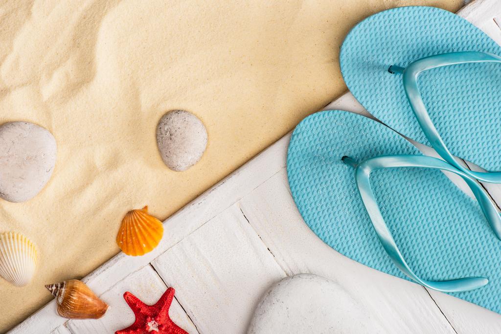 Top view of seashells near starfish and blue flip flops on white wooden planks and sand - Photo, Image