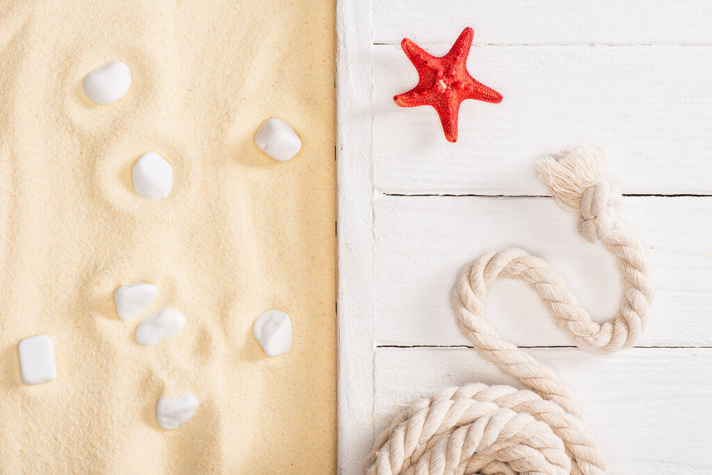 Vista superior de estrelas-do-mar vermelhas e corda em tábuas de madeira branca e pedras marinhas na areia
 - Foto, Imagem