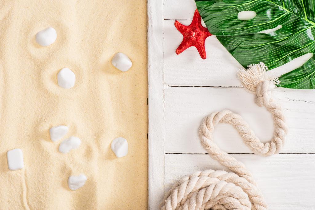 Верхний вид морской звезды, веревки и зеленого листа на белых деревянных досках возле гальки на песке
 - Фото, изображение