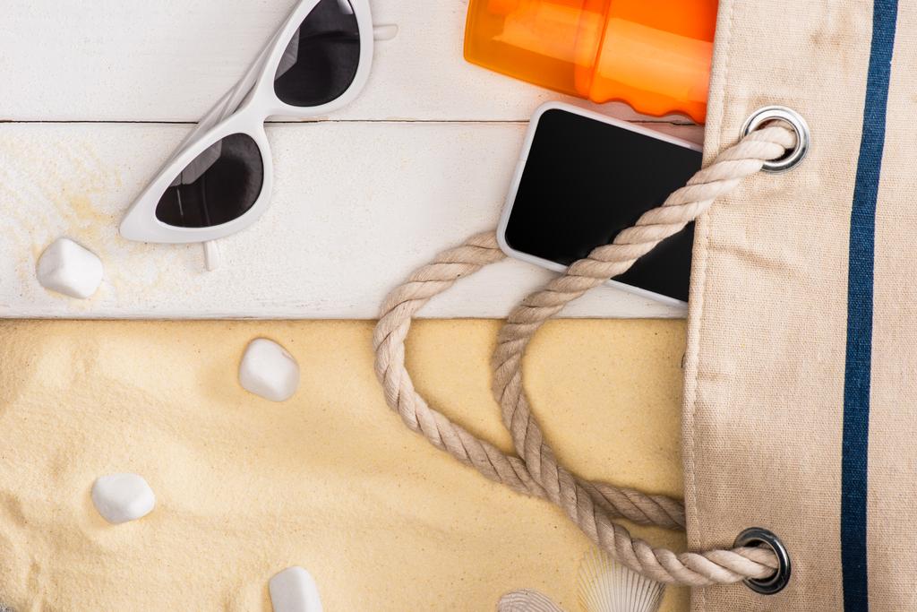 Bovenaanzicht van smartphone en zonnebrandcrème nabij tas en zonnebril op witte houten planken en zeestenen op zand - Foto, afbeelding