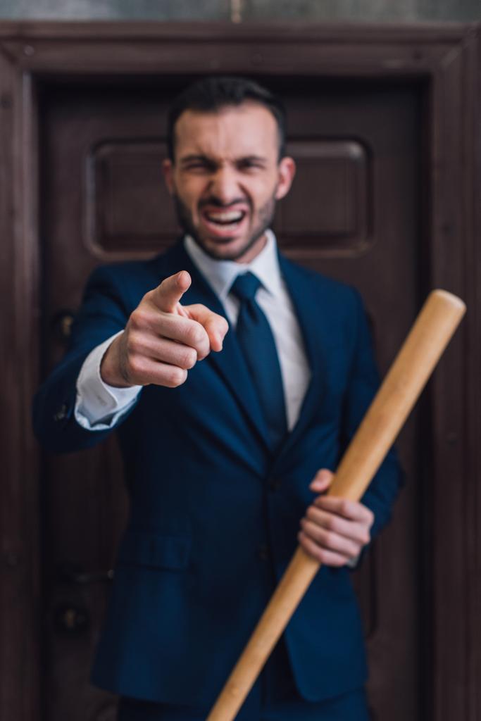 Selectieve focus van boze verzamelaar met honkbalknuppel wijzend met vinger en schreeuwend naast de deur in de kamer - Foto, afbeelding