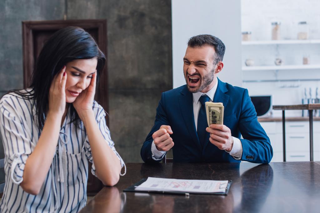 Opgewonden verzamelaar met dollarbiljetten in de buurt van gestresste vrouw aan tafel in de kamer - Foto, afbeelding