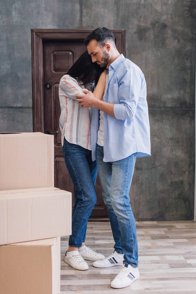 Мужчина обнимает расстроенную жену возле коробок в комнате
 - Фото, изображение