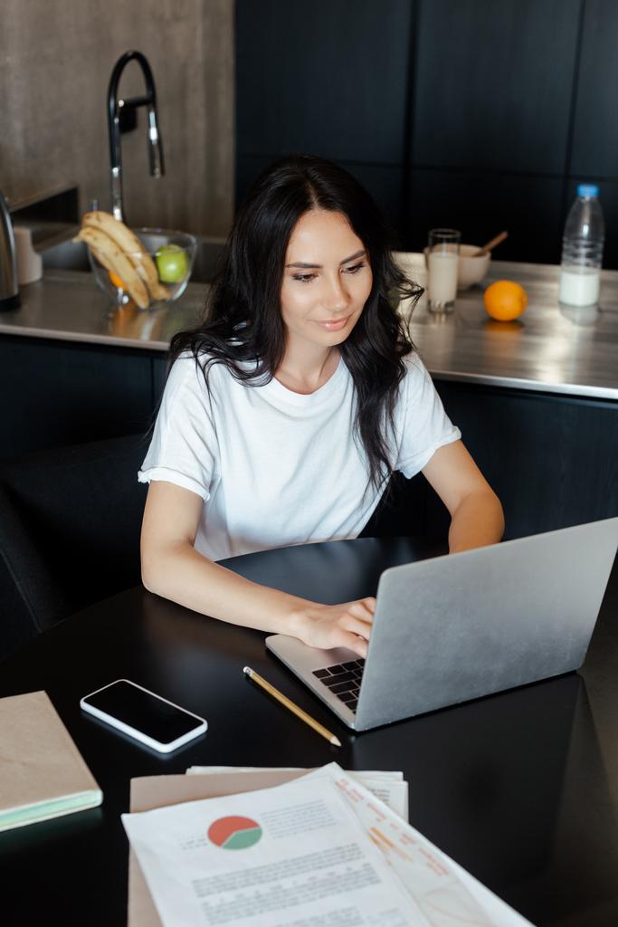 красивая женщина, работающая с ноутбуком, смартфоном и бизнес-документами на кухне по самоизоляции
 - Фото, изображение