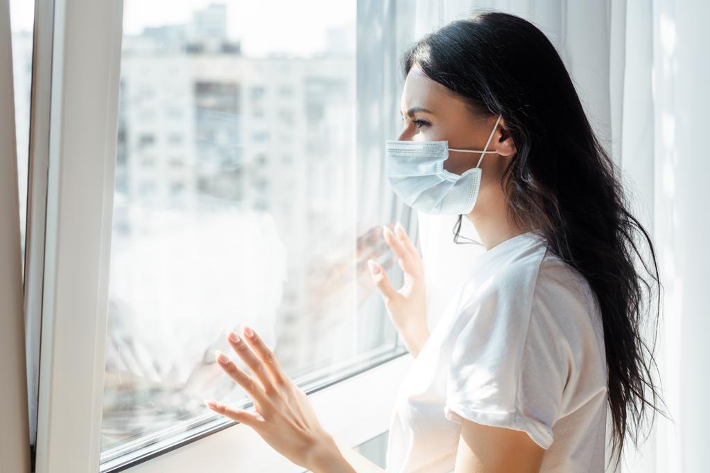 больная молодая женщина в медицинской маске смотрит в окно на самоизоляцию
 - Фото, изображение