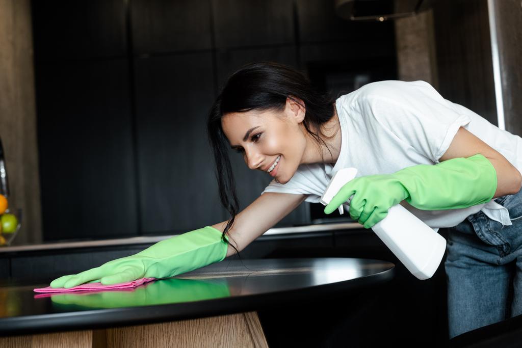 gelukkig aantrekkelijk vrouw in latex handschoenen reinigen tafel met roze doek en spray fles - Foto, afbeelding