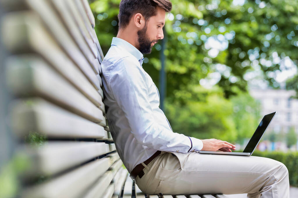 Porträt eines attraktiven Geschäftsmannes, der auf einer Bank sitzt, während er an seinem Laptop arbeitet - Foto, Bild