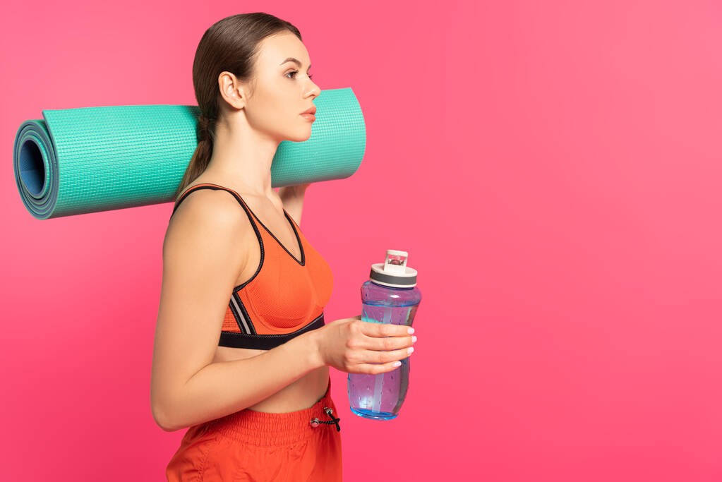 ελκυστική αθλήτρια κρατώντας αθλητικό μπουκάλι με νερό και στρώμα γυμναστικής που απομονώνονται σε ροζ  - Φωτογραφία, εικόνα