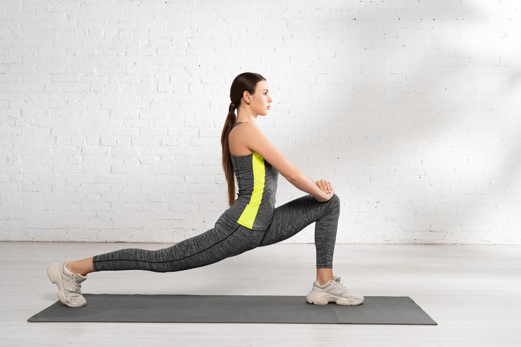 side view of flexible sportswoman exercising on fitness mat near white brick wall - Photo, Image