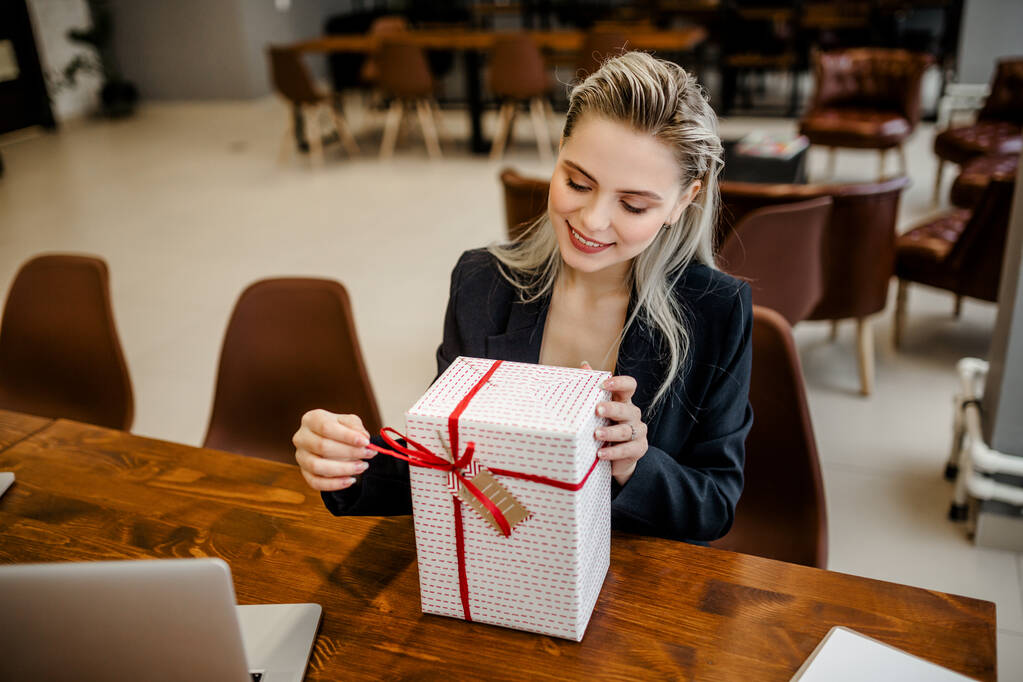 Piękna blondynka bizneswoman siedzi przy biurku w biurze i trzyma prezent, który dostali koledzy z pracy - Zdjęcie, obraz