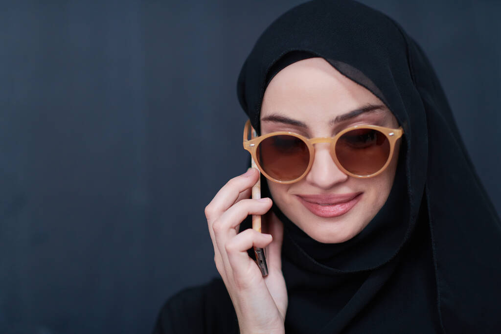 νεαρή μοντέρνα μουσουλμάνα επιχειρηματίας χρησιμοποιώντας smartphone φορώντας γυαλιά ηλίου και χιτζάμπ ρούχα μπροστά από μαύρο μαυροπίνακα - Φωτογραφία, εικόνα