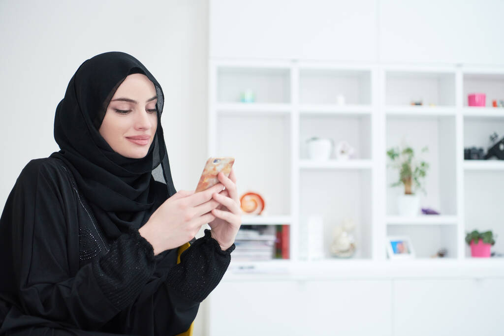 Молода сучасна мусульманська бізнес-леді використовує смартфон в традиційному одязі хіджабу вдома
 - Фото, зображення