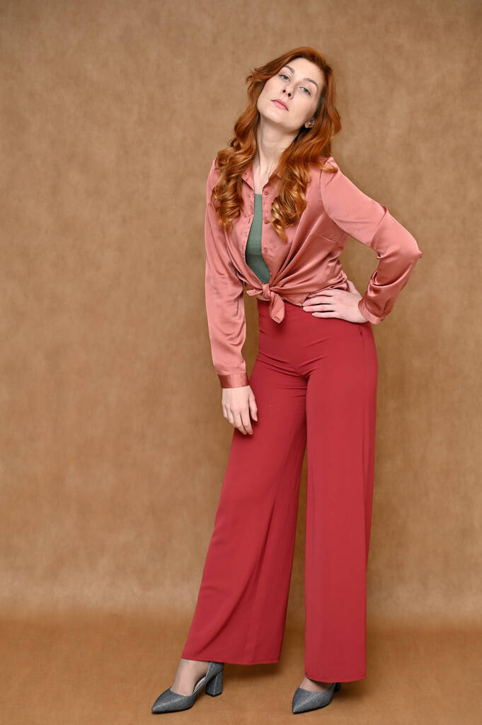 In voller Länge vertikales Porträt einer posierenden gerade kaukasischen roten Haare nette junge Frau auf einem beigen Hintergrund. Das Model ist bekleidet mit einem rosa Hemd und einer roten Hose, grauen Schuhen. - Foto, Bild