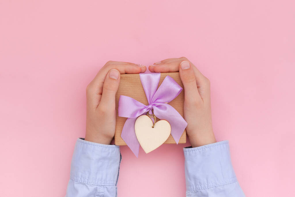 Kid's hands holding gift box wrapped in craft paper and tied with bow on pink background. Concept Mother's Day or Birthday. Greeting card. - Photo, Image