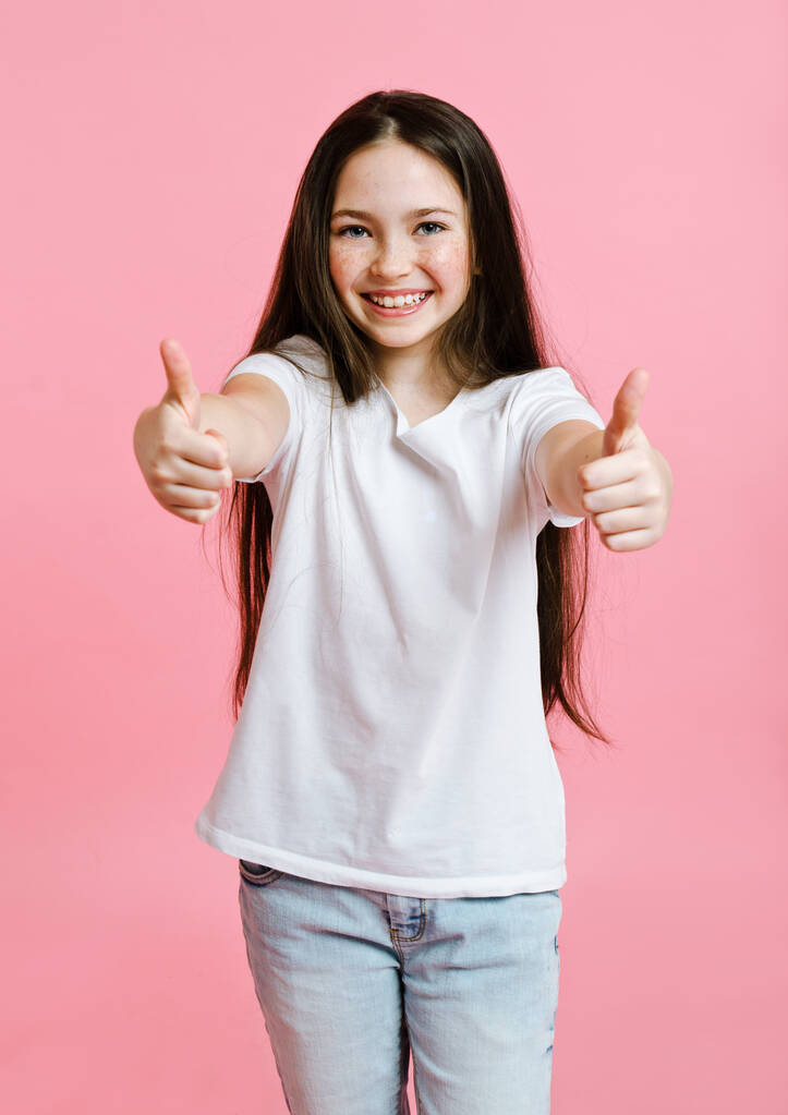 Portret uroczej, uśmiechniętej dziewczynki nastolatki w białej koszulce z dwoma palcami na różowym tle - Zdjęcie, obraz