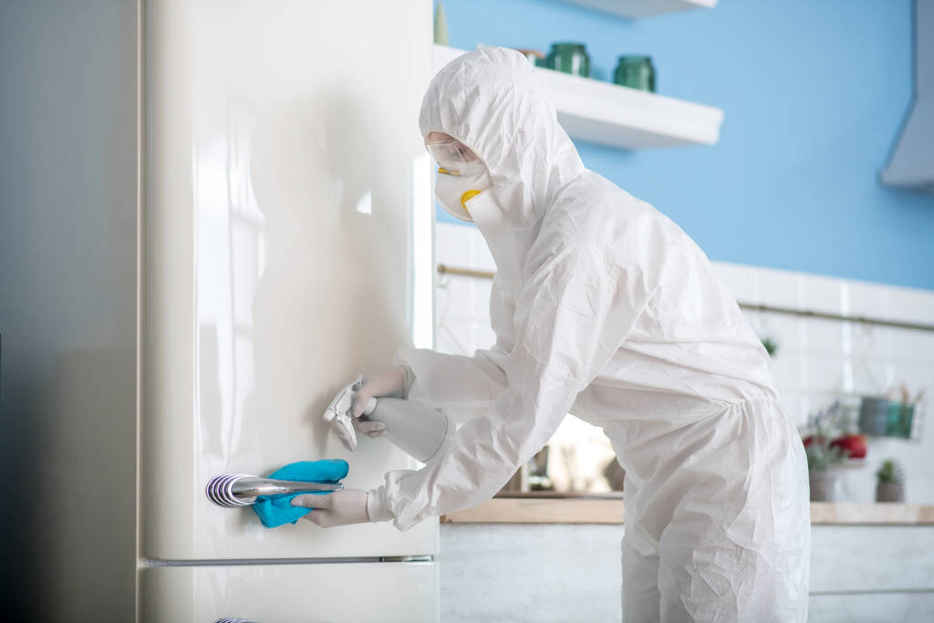 Mujer en ropa de trabajo blanca y respirador desinfectando la nevera
 - Foto, imagen