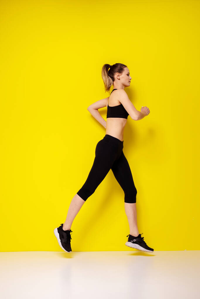 Sarı arka planda siluetli bir kadın koşucu. Çekici bir kadının şık spor elbiseli fotoğrafı. Dinamik hareket. Yan görüş. Spor ve sağlıklı yaşam tarzı - Fotoğraf, Görsel