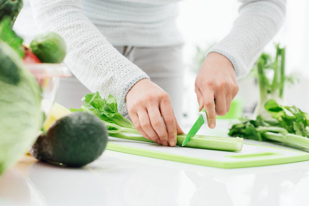 Woman preparing food in her kitchen, she is chopping fresh celery on a cutting board with a knife - Photo, Image