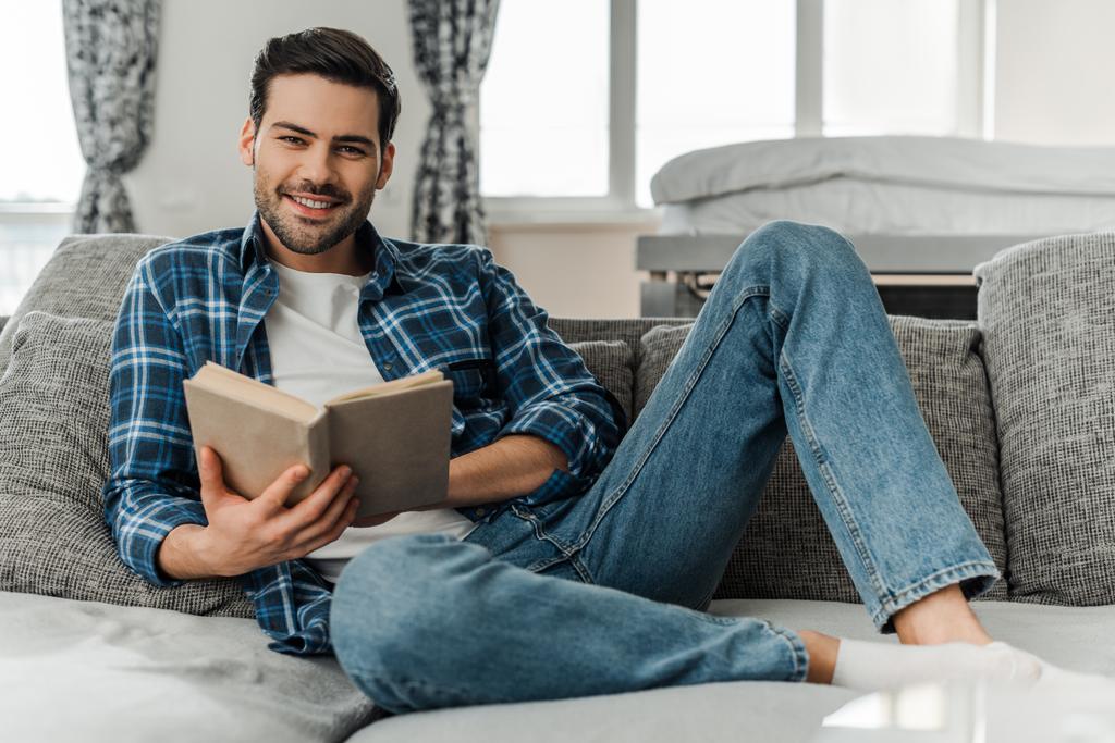 Selektiver Fokus eines Mannes, der in die Kamera lächelt, während er sein Buch zu Hause auf dem Sofa hält - Foto, Bild