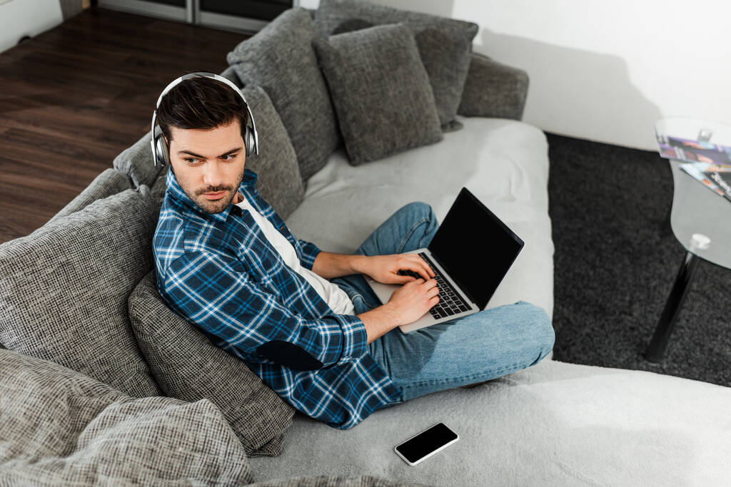 Visão de alto ângulo do homem em fones de ouvido usando laptop perto de smartphone no sofá
  - Foto, Imagem