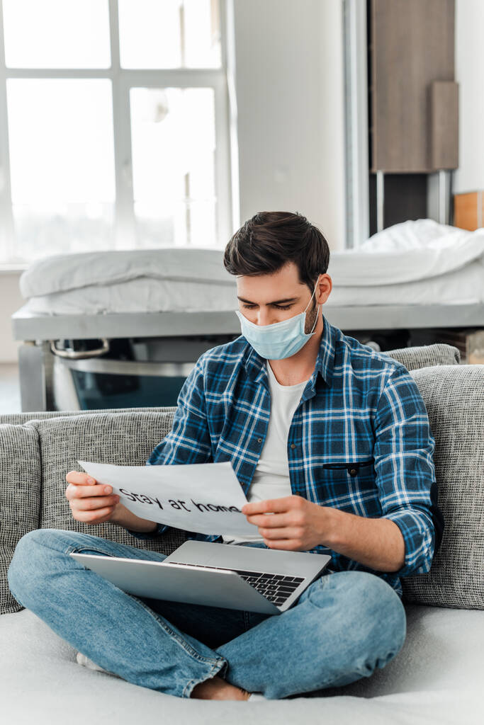 Фрілансер в медичній масці тримає карту з перебуванням вдома написанням і ноутбуком на дивані
  - Фото, зображення