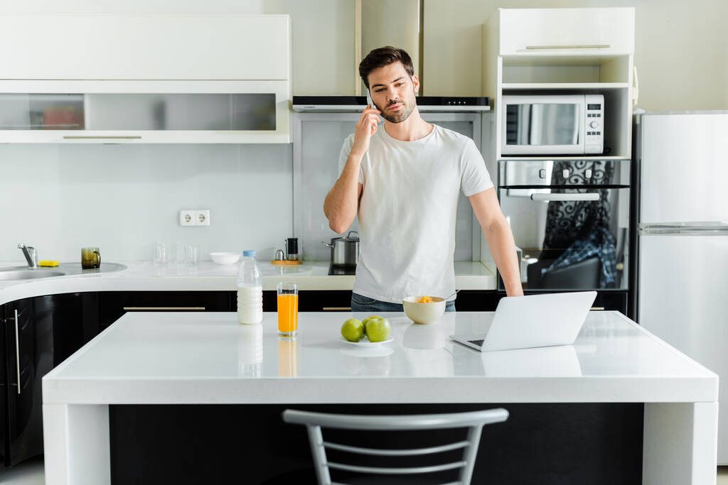 Bilgisayarın yanında akıllı telefondan konuşan adam, mısır gevreği ve portakal suyu mutfak masasında.  - Fotoğraf, Görsel