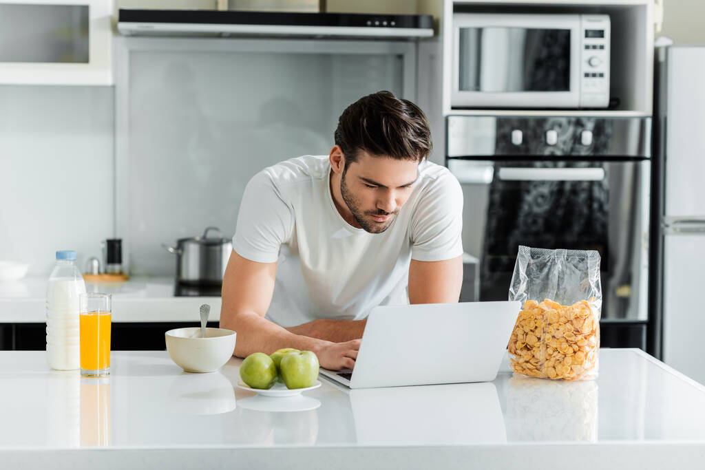 Homem usando laptop perto de cereais e suco de laranja na mesa da cozinha
  - Foto, Imagem