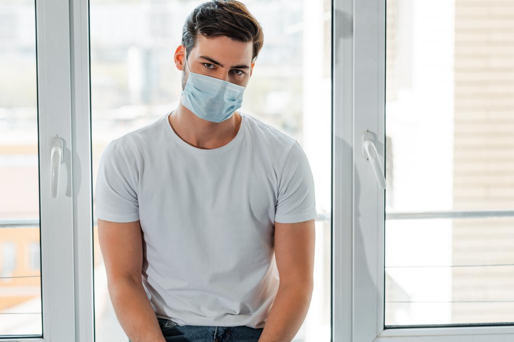Hombre con máscara médica mirando a la cámara cerca de la ventana en casa
  - Foto, imagen