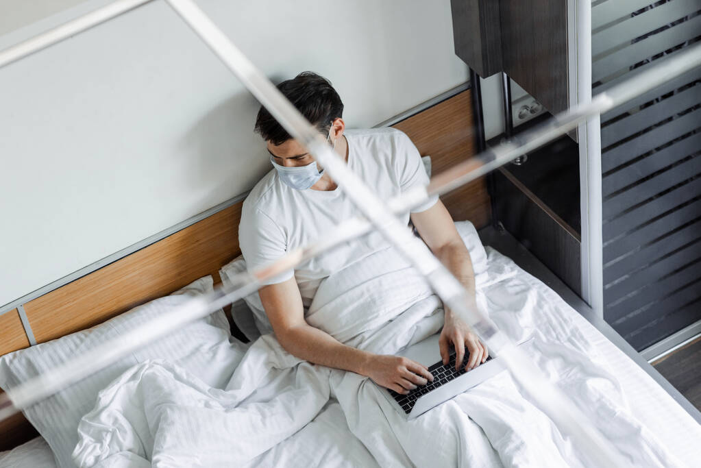 Pohled shora na muže v lékařské masce pomocí notebooku na posteli  - Fotografie, Obrázek