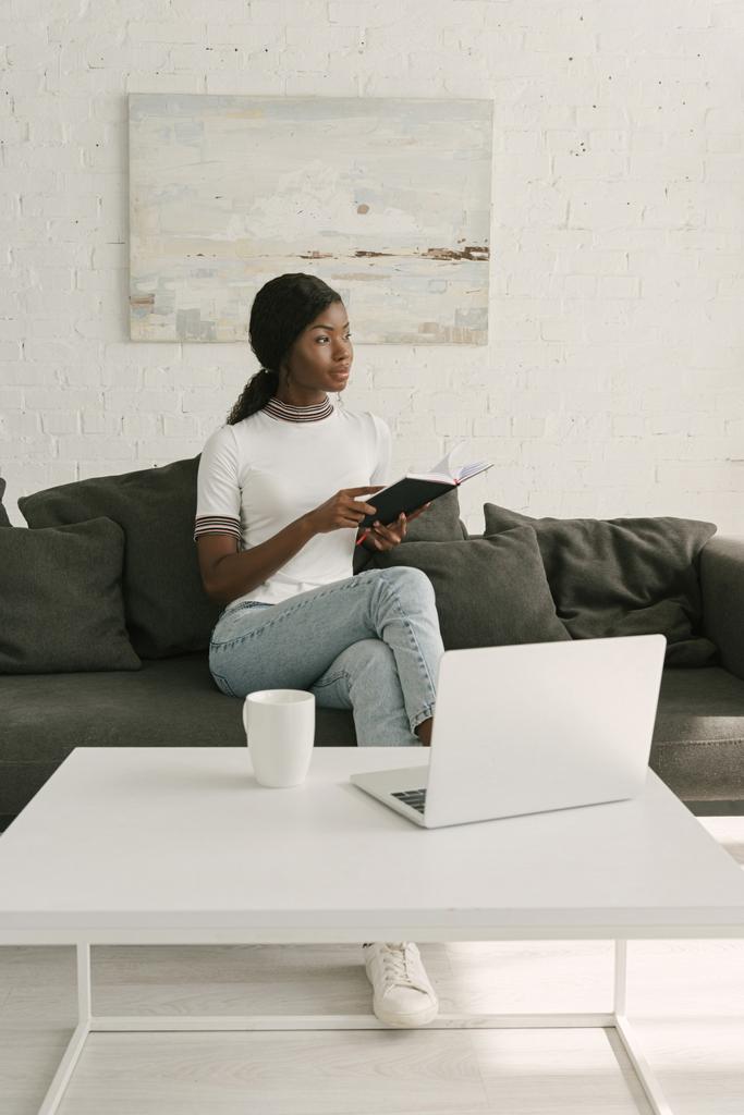 thoughtful african american freelancer holding notebook while sitting on sofa near table with laptop - Photo, Image