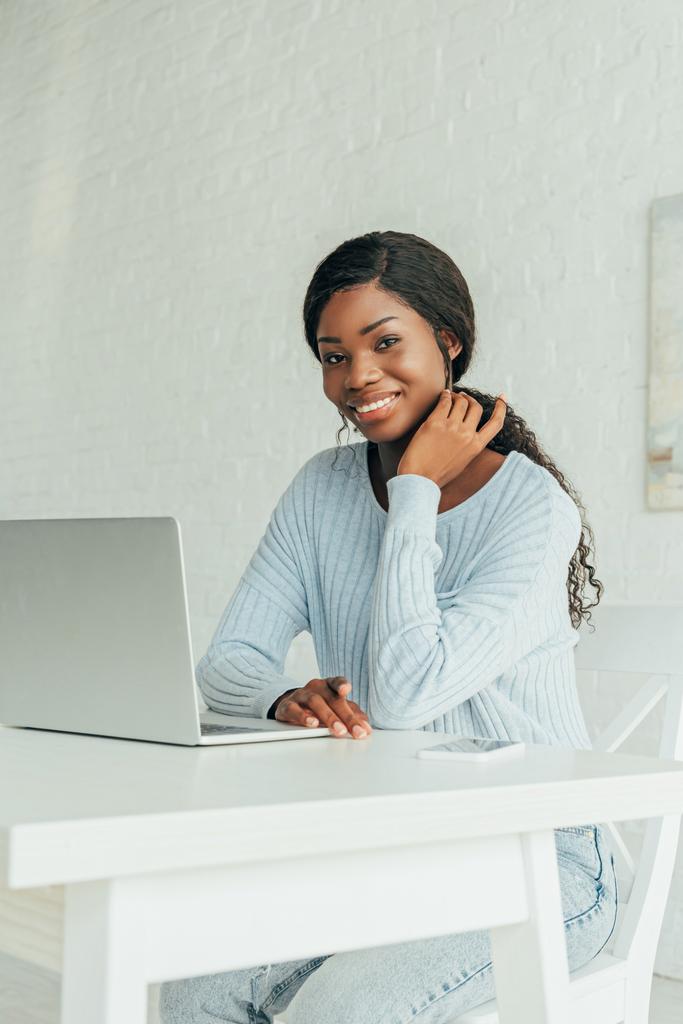 счастливый африканский американский фрилансер улыбается в камеру, сидя рядом с ноутбуком дома
 - Фото, изображение