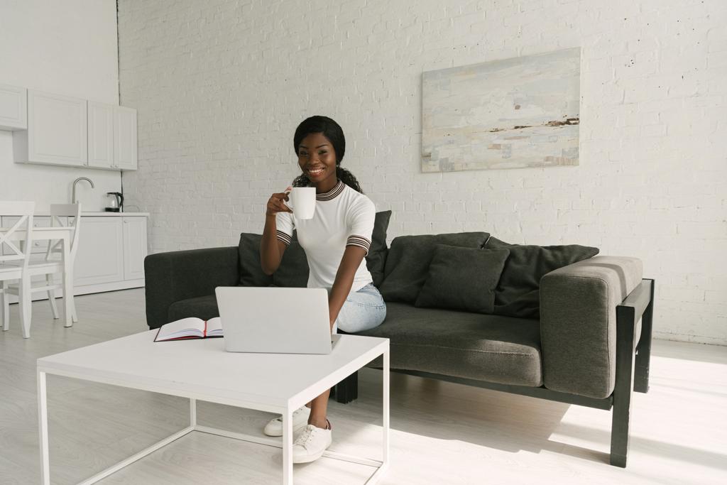 счастливый африканский американский фрилансер улыбается в камеру, сидя на диване с чашкой кофе рядом со столом с ноутбуком
 - Фото, изображение