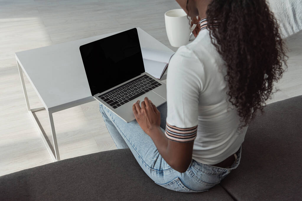 vysoký úhel pohledu africké americké dívky pomocí notebooku s prázdnou obrazovkou a drží šálek kávy - Fotografie, Obrázek