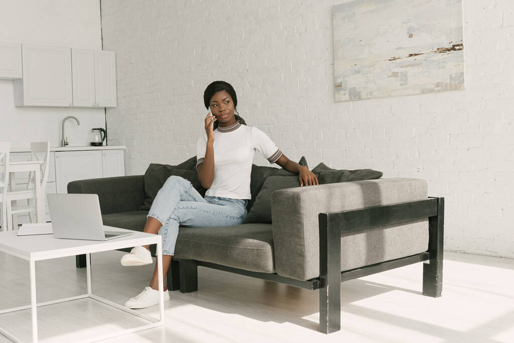 nachdenklicher afrikanisch-amerikanischer Freelancer, der auf dem Sofa am Tisch mit Laptop sitzt und auf dem Smartphone spricht  - Foto, Bild