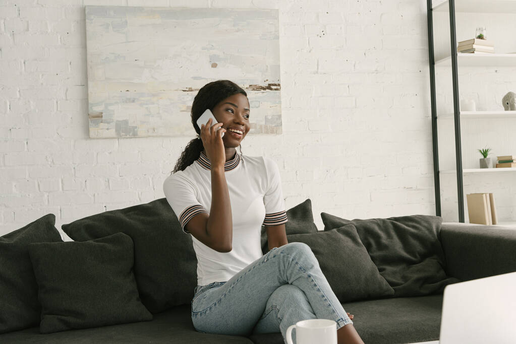 fröhliches afrikanisch-amerikanisches Mädchen sitzt auf dem Sofa, spricht auf dem Smartphone und schaut weg - Foto, Bild