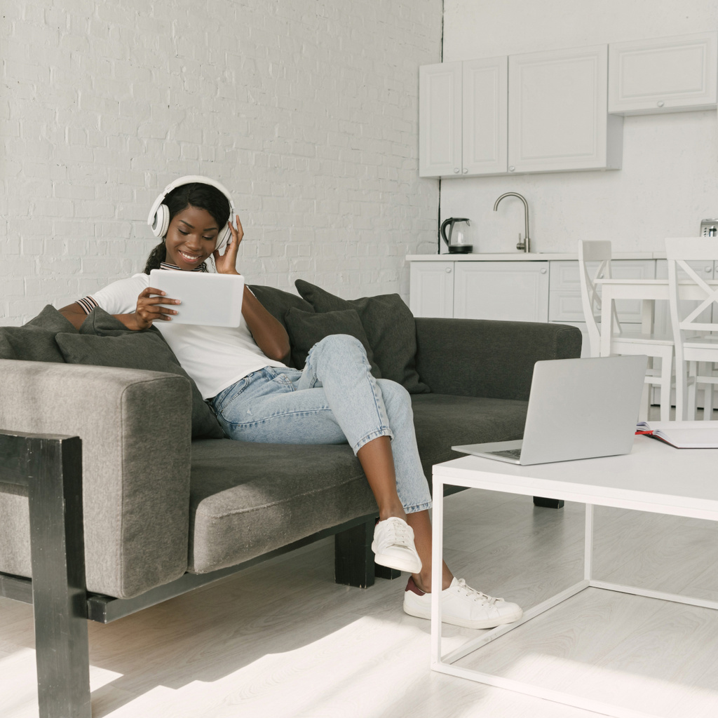 ノートパソコンとテーブルの近くのソファに座っている間、デジタルタブレットを使用してワイヤレスヘッドフォンで幸せなアフリカ系アメリカ人フリーランサー - 写真・画像