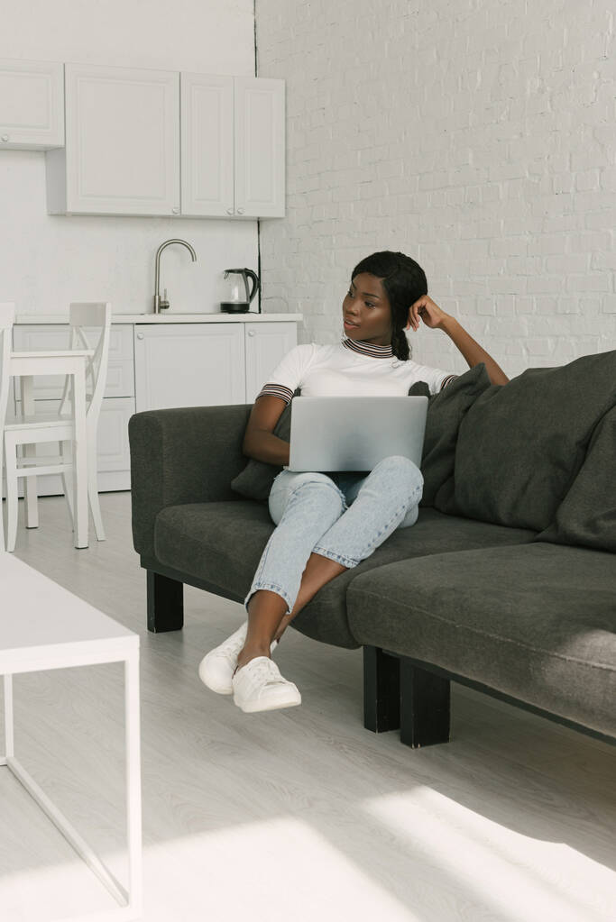 freelancer afro-americano pensativo sentado no sofá com laptop e olhando para longe
 - Foto, Imagem
